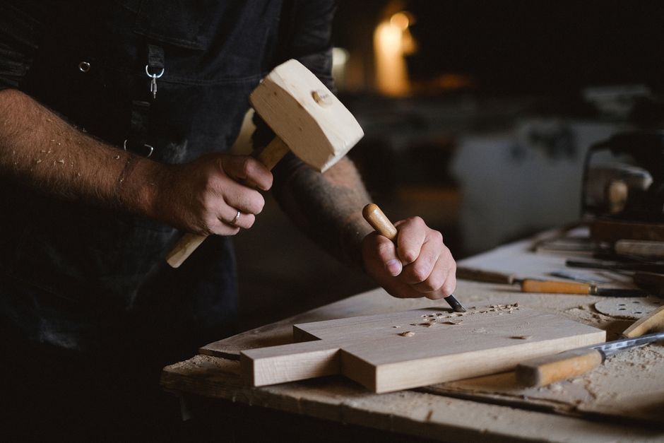 Unrecognizable male artisan using wooden hammer and chisel to carve lumber plank while working at table with special instruments on blurred background